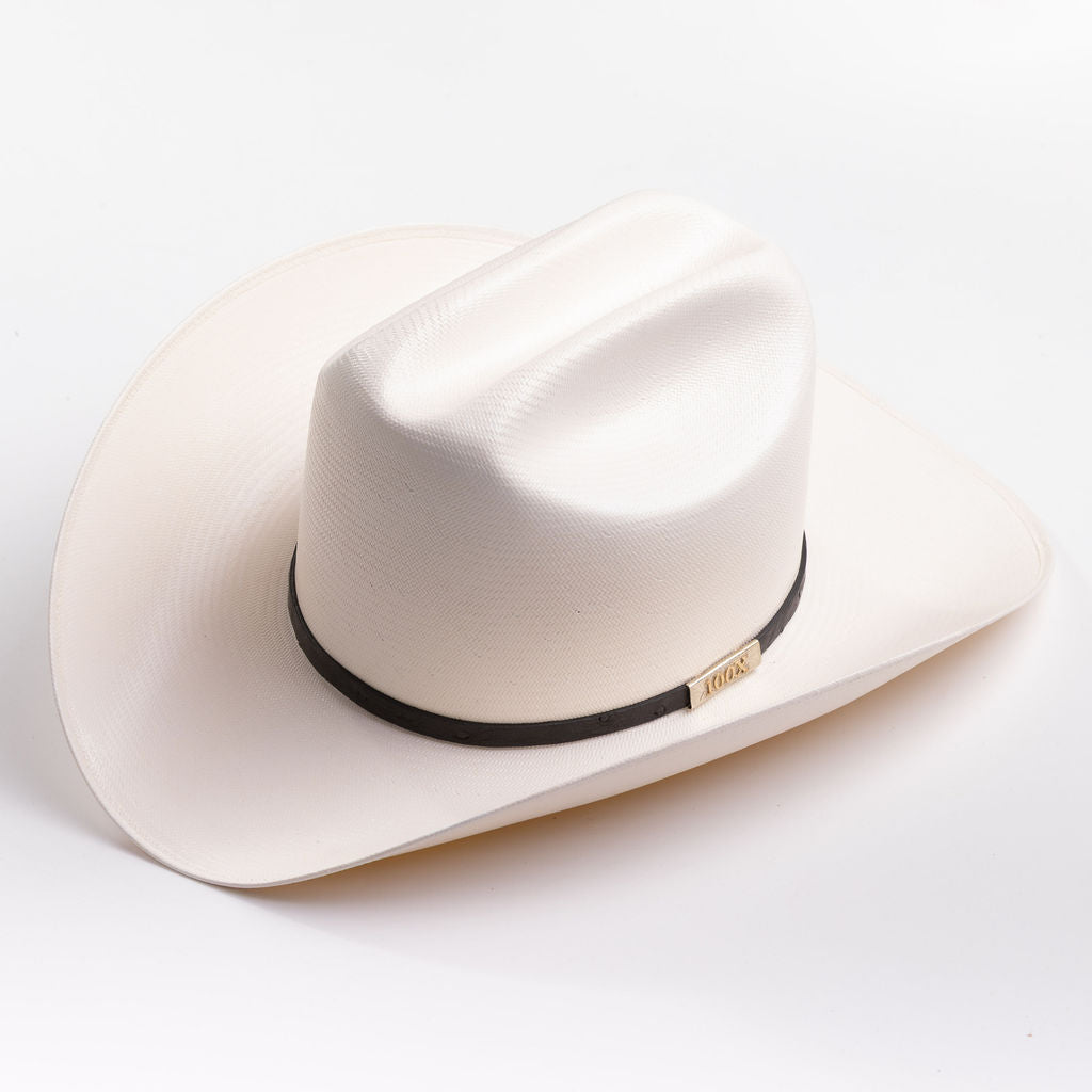 A cream-colored cowboy hat with a black band and a small gold metal logo tag, photographed on a clean light gray background