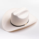 A cream-colored cowboy hat with a black band and a small gold metal logo tag, photographed on a clean light gray background