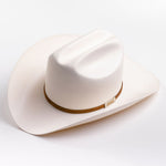 A cream-colored cowboy hat with a cognac band and a small gold metal logo tag, photographed on a clean light gray background