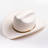 A cream-colored cowboy hat with a cognac band and a small gold metal logo tag, photographed on a clean light gray background