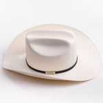 A cream-colored cowboy hat with a black band and a small gold metal logo tag, photographed on a clean light gray background