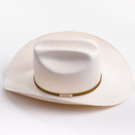 A cream-colored cowboy hat with a cognac band and a small gold metal logo tag, photographed on a clean light gray background