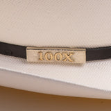 Close-up of a white woven hat with a black band and a rectangular gold-tone metal plaque engraved with "100X"
