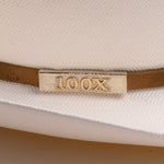 Close-up of a white woven hat with a cognac band and a rectangular gold-tone metal plaque engraved with "100X"