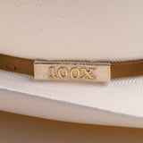 Close-up of a white woven hat with a cognac band and a rectangular gold-tone metal plaque engraved with "100X"
