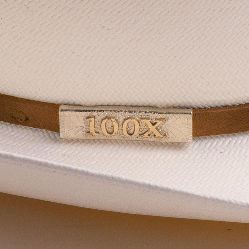 Close-up of a white woven hat with a cognac band and a rectangular gold-tone metal plaque engraved with "100X"