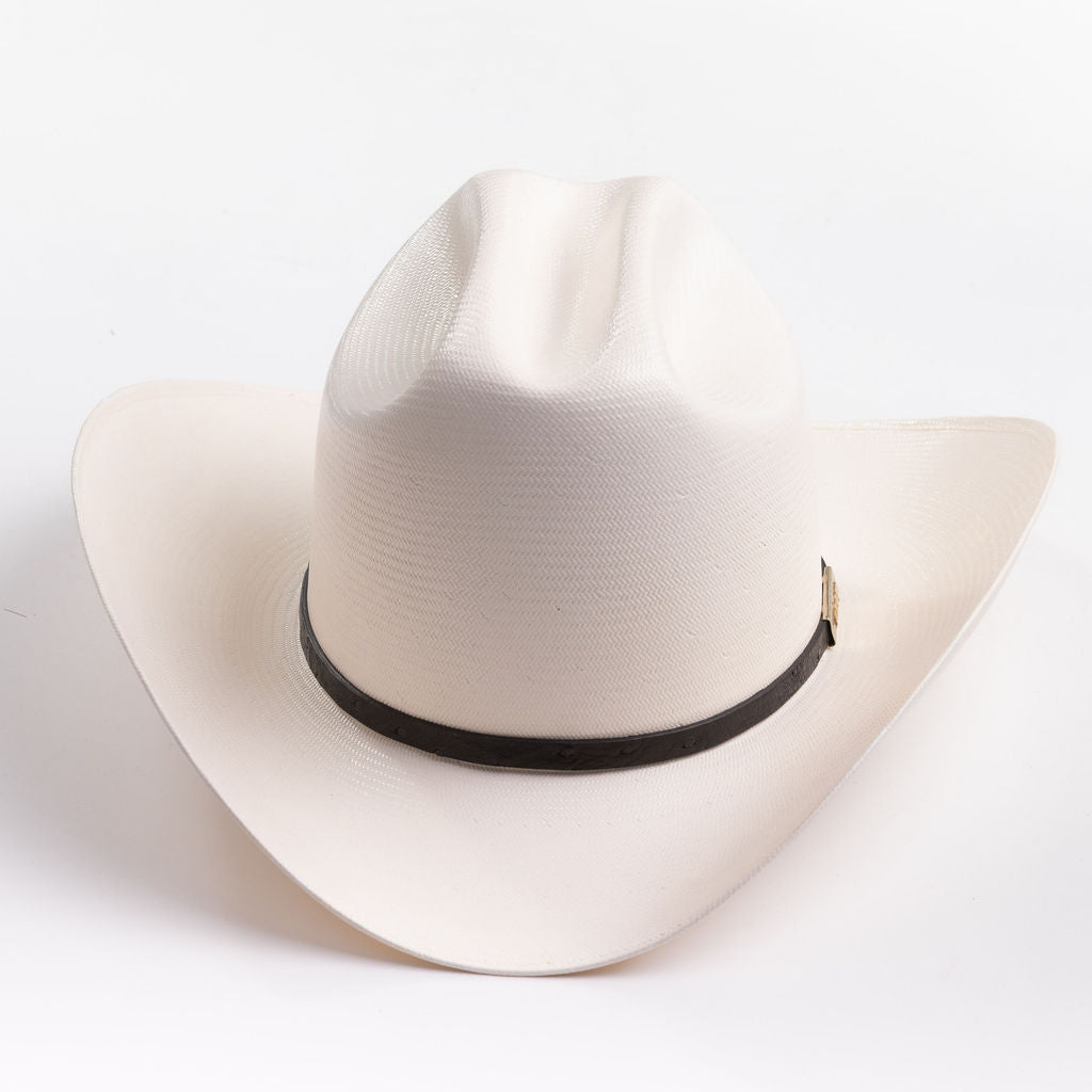 A cream-colored cowboy hat with a black band and a small gold metal logo tag, photographed on a clean light gray background