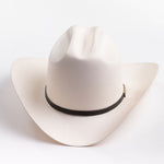 A cream-colored cowboy hat with a black band and a small gold metal logo tag, photographed on a clean light gray background