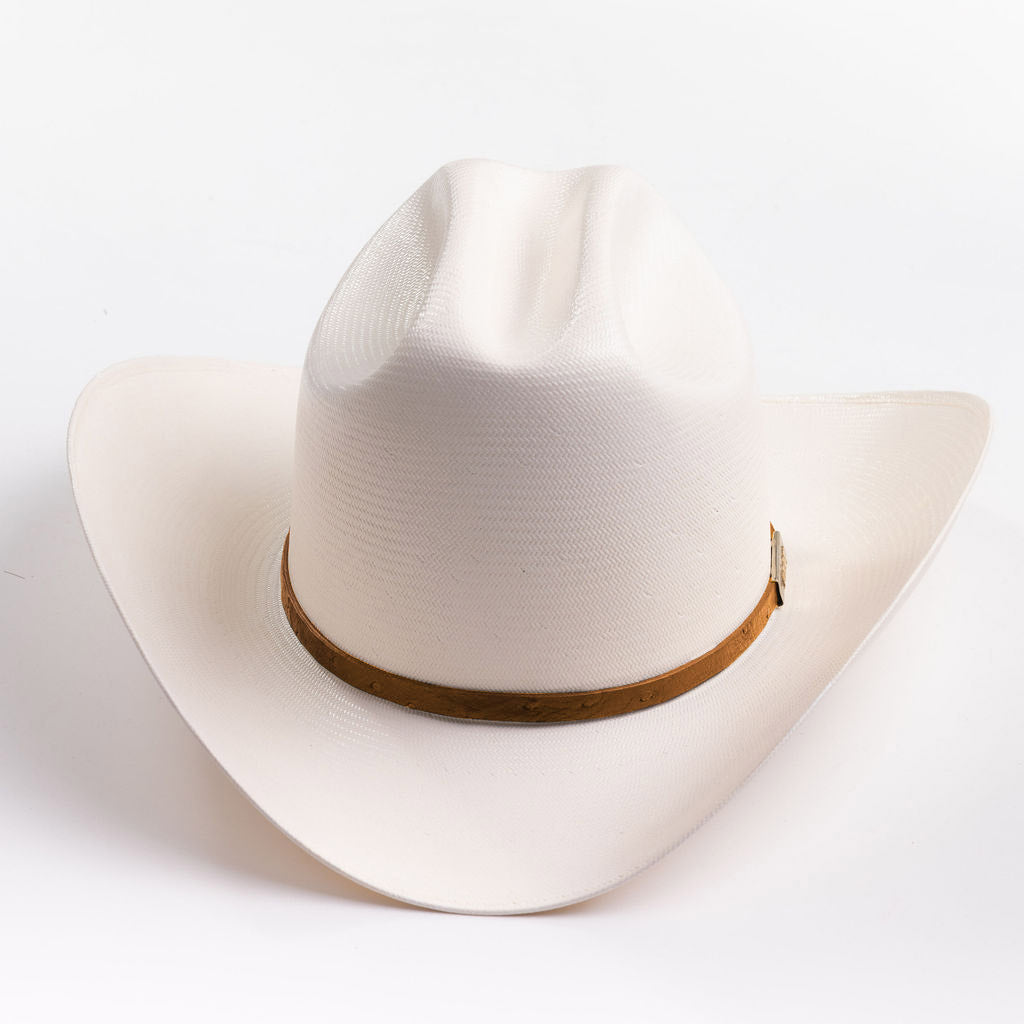 A cream-colored cowboy hat with a cognac band and a small gold metal logo tag, photographed on a clean light gray background