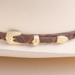 A close-up of a brown leather hat band with ornate gold-toned conchos, shown resting on a light cream-colored hat
