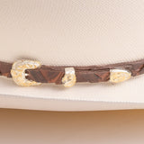 A close-up of a brown leather hat band with ornate gold-toned conchos, shown resting on a light cream-colored hat
