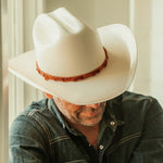 Man wearing an off-white cowboy hat with buttercup colored band with his head down with his eyes not visible while also wearing a dark denim shirt