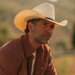 Man wearing an off-white colored cowboy hat and red button-up shirt against a grassy blurred background