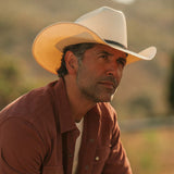 Man wearing an off-white colored cowboy hat and red button-up shirt against a grassy blurred background