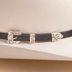 A close-up of a black leather hat band with ornate silver-toned metal buckles and decorative end pieces, shown on a white textured cowboy hat