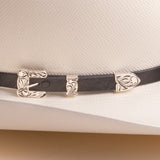 A close-up of a black leather hat band with ornate silver-toned metal buckles and decorative end pieces, shown on a white textured cowboy hat