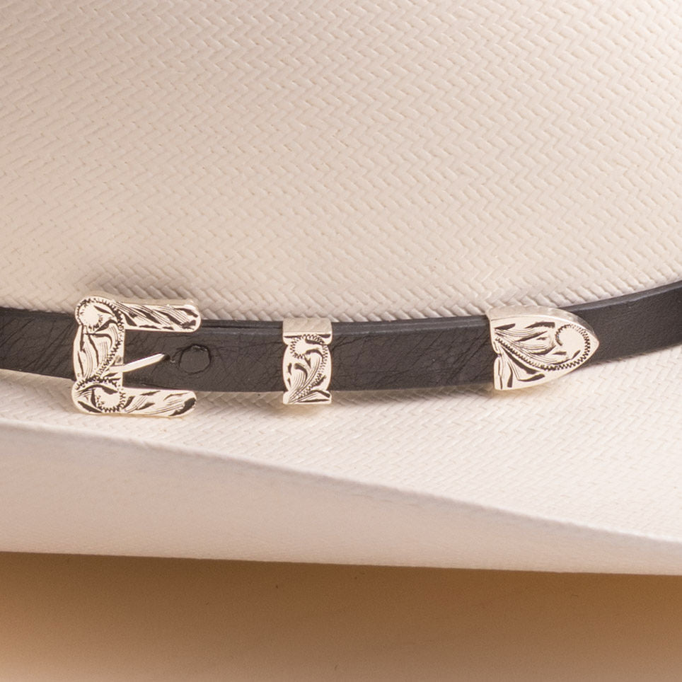 A close-up of a black leather hat band with ornate silver-toned metal buckles and decorative end pieces, shown on a white textured cowboy hat