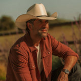 Man wearing an off-white colored cowboy hat and red button-up shirt against a grassy fenced off blurred background