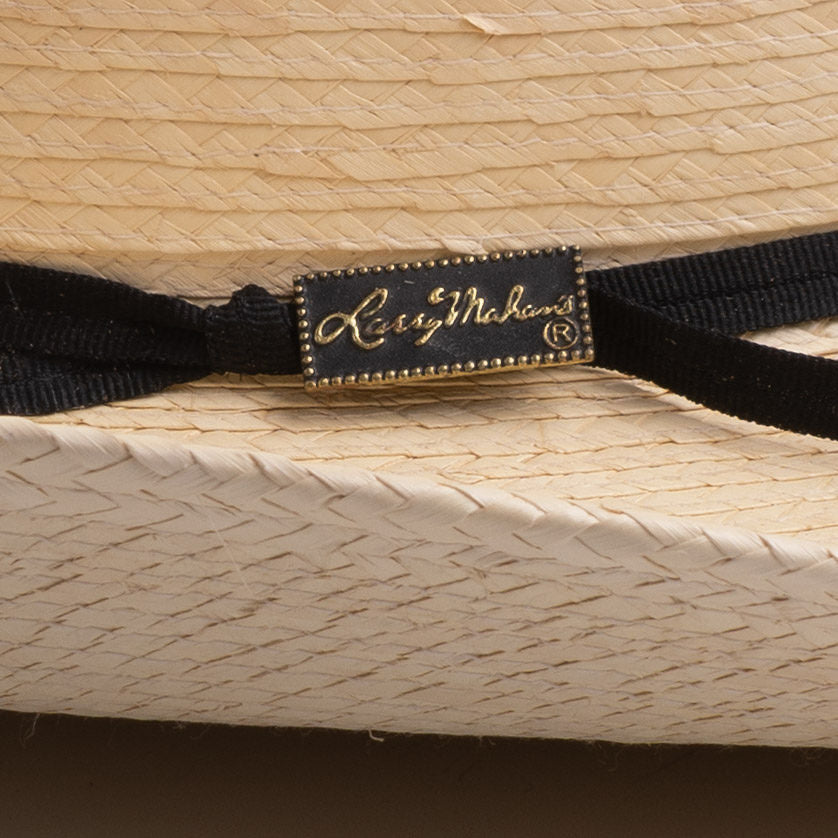 A close-up of a black ribbon hatband with a rectangular black label featuring gold cursive script that reads 'Larry Mahan' and a gold border, shown tied around a woven straw hat