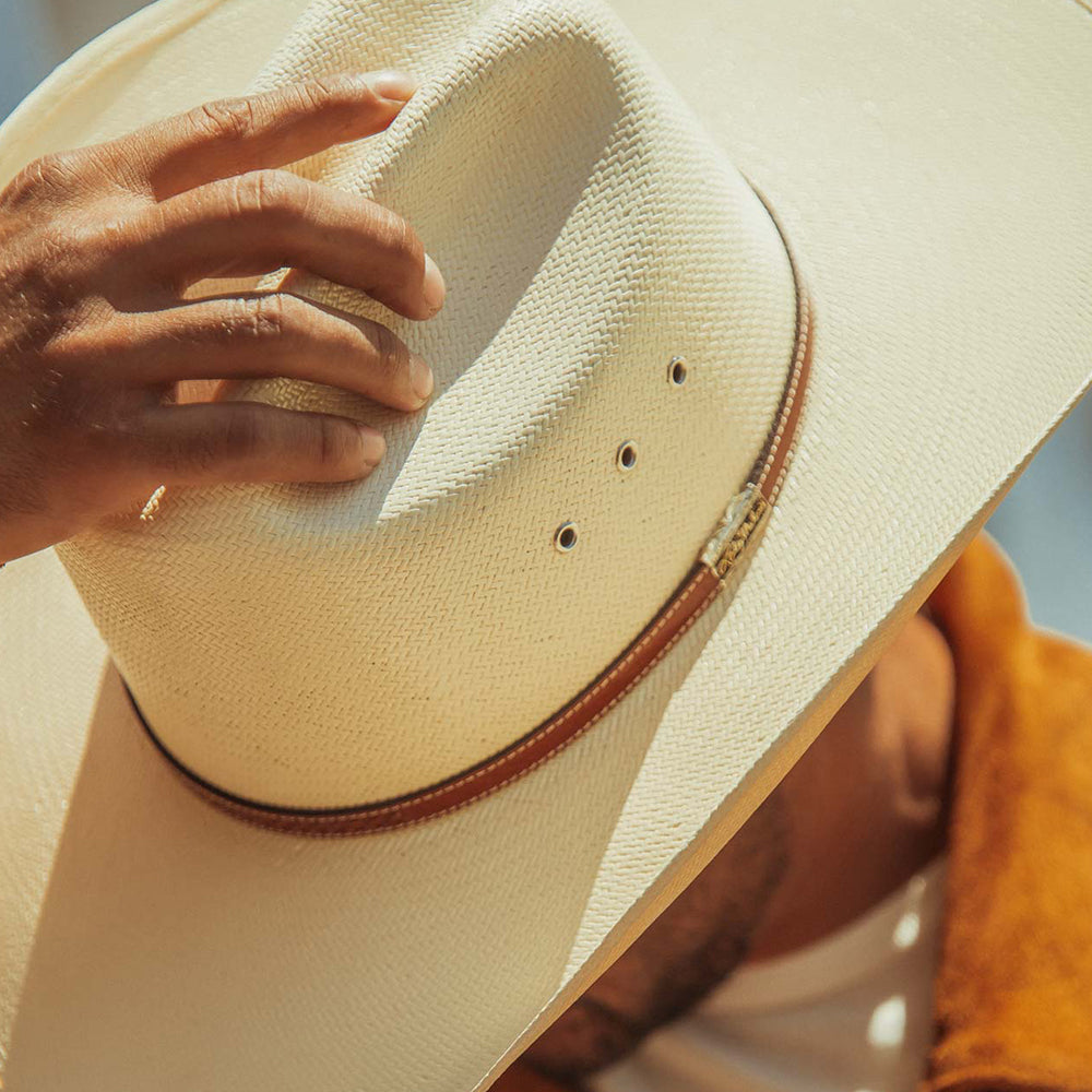 A close-up of a cream-colored woven cowboy hat with a brown band, held by a hand. The hat features three ventilation eyelets and is shown in warm, natural light