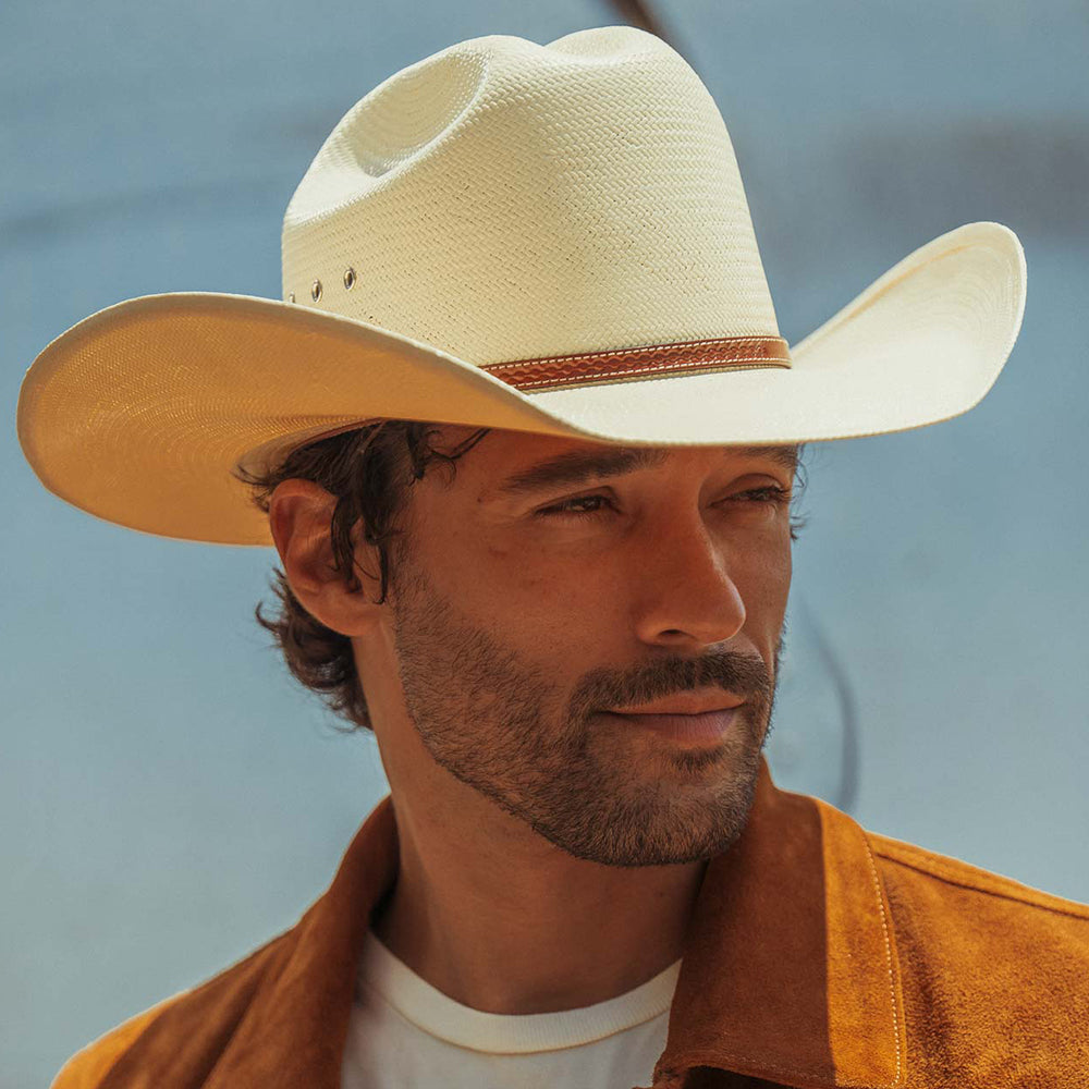 A close-up portrait of a man wearing a cream-colored woven cowboy hat with a brown band and a brown suede jacket against a soft blue-gray background