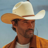 A close-up portrait of a man wearing a cream-colored woven cowboy hat with a brown band and a brown suede jacket against a soft blue-gray background