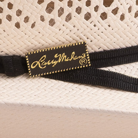 Close-up of a black woven hat band with a rectangular black label featuring gold script reading 'Larry Mahan's' and gold beaded edges, set against an off-white woven straw hat
