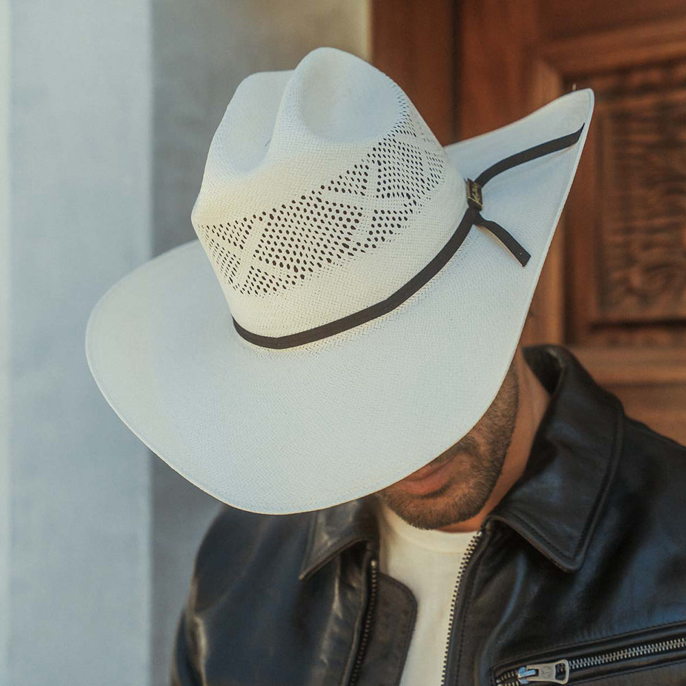 A man wearing a white cowboy hat with a woven pattern and a black band, paired with a black leather jacket and white t-shirt, set against a rustic wooden door background