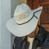 A man wearing a white cowboy hat with a woven pattern and a black band, paired with a black leather jacket and white t-shirt, set against a rustic wooden door background