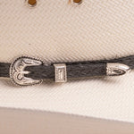 A close-up of a black braided leather hat strap with ornate silver-toned metal band, resting on a textured off-white woven surface of the hat