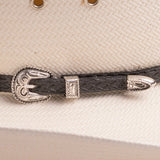 A close-up of a black braided leather hat strap with ornate silver-toned metal band, resting on a textured off-white woven surface of the hat
