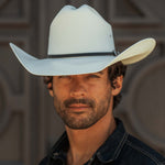 A man wearing a white cowboy hat with a black band and a dark denim shirt, shown in a close-up portrait