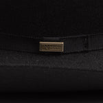 Close-up of a black felt hat with a black ribbon band and a small gold-tone metal pin engraved with "Worn by Legends"