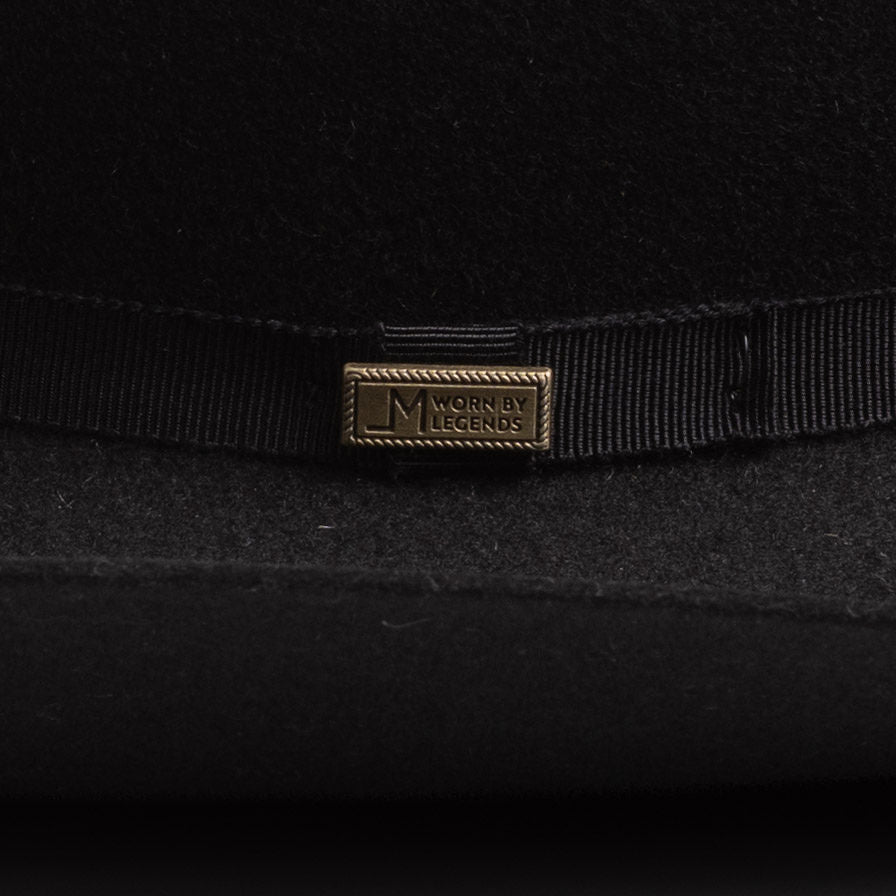 Close-up of a black felt hat with a black ribbon band and a small gold-tone metal pin engraved with "Worn by Legends"