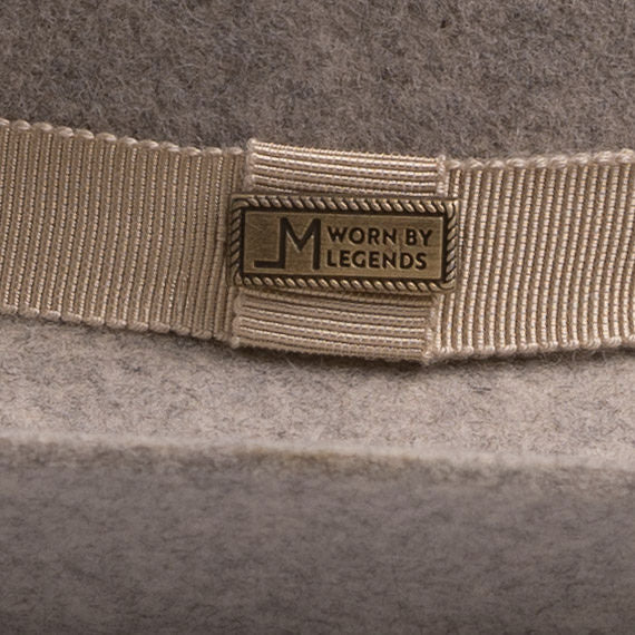 Close-up of a beige grosgrain ribbon band with a rectangular metal logo plate reading "Worn by Legends" on a gray felt hat