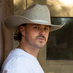 A man wearing a natural colored felt cowboy hat and a white t-shirt, looking over his shoulder in a rustic setting.