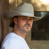 A man wearing a natural colored felt cowboy hat and a white t-shirt, looking over his shoulder in a rustic setting.