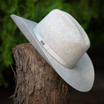 A natural colored felt wide-brim hat with a white band and small silver logo plaque, resting on a textured tree stump with a blurred green outdoor background.