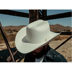 Bearded man wearing a 6X Real white cowboy hat looking downwards while underneath a tall wooden windmill