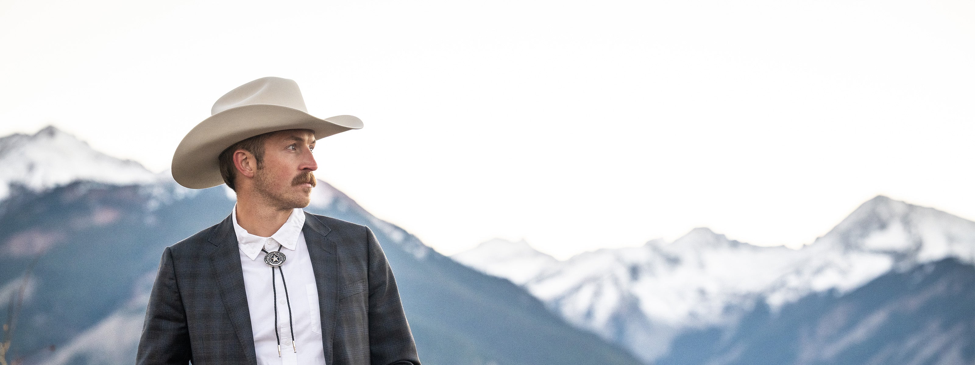 Man wearing a cowboy hat and plaid jacket with mountains in the background