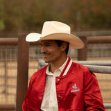 Smiling man wearing a 10X Brindle brown cowboy hat with a bright red jacket and white under shirt standing near metal fences