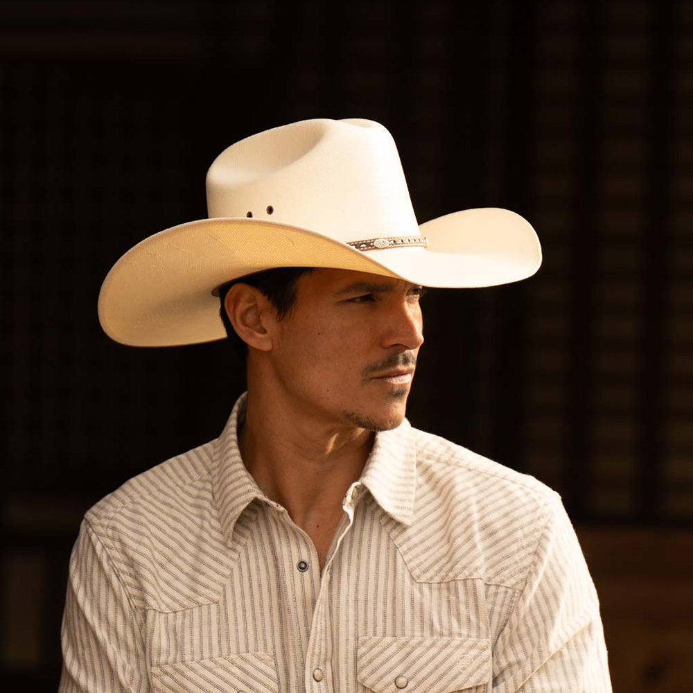 Man wearing a 10X Bull ivory cowboy hat with white striped long sleeve shirt standing against a dark brown background