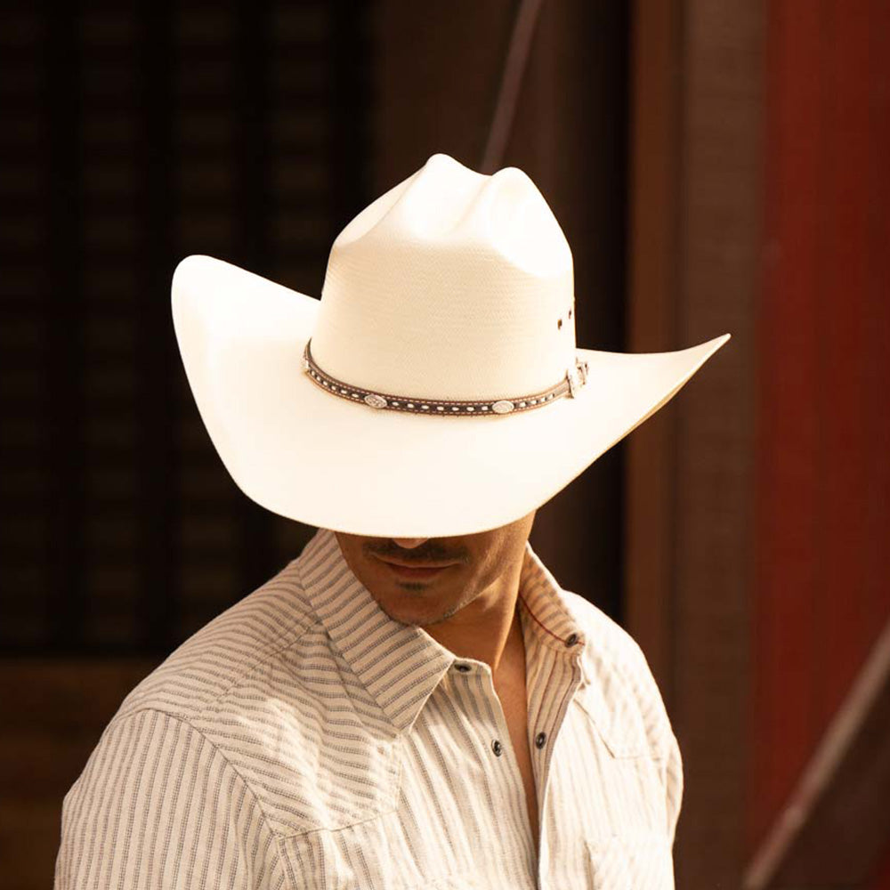 Man wearing a 10X Bull ivory cowboy hat with white striped long sleeve shirt standing against a dark brown and red background
