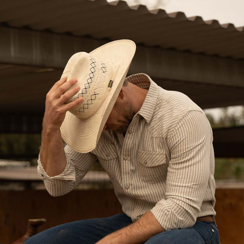 Bearded man wearing a 10X Cowboy Cruz tan and ivory cowboy hat with plaid white shirt and blue jeans looking down while facing left with his right hand on the top of his hat