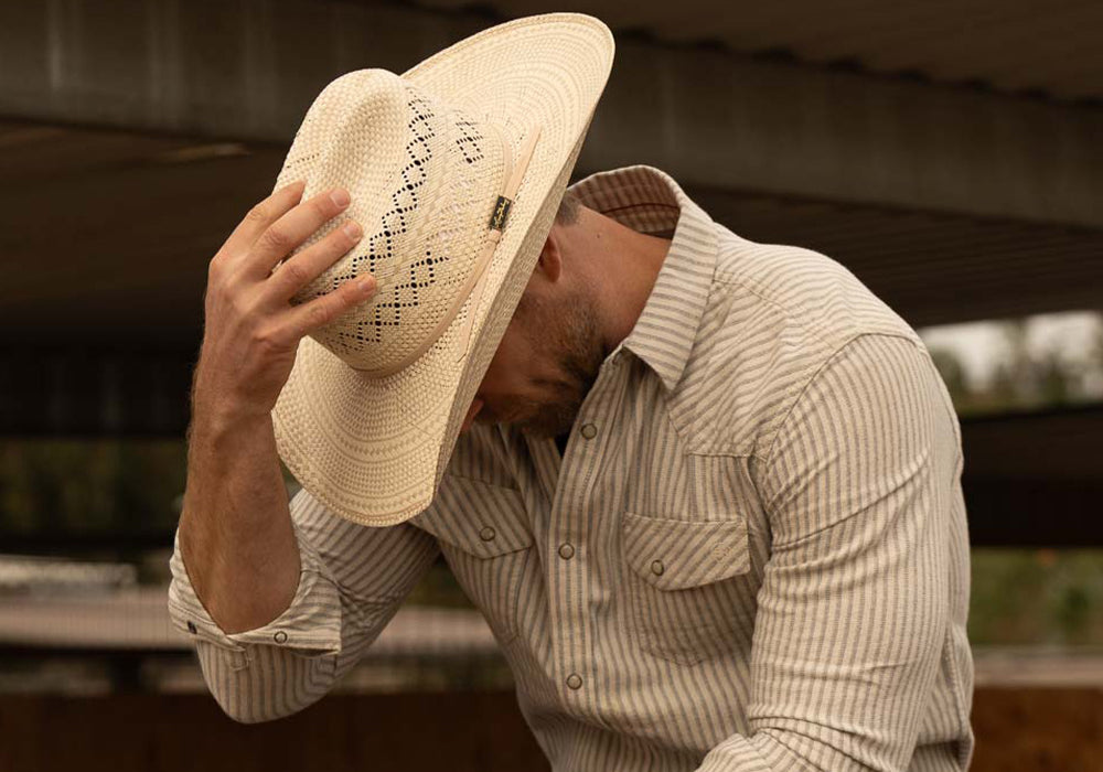 Bearded man wearing a 10X Cowboy Cruz tan and ivory cowboy hat with plaid white shirt and blue jeans looking down while facing left with his right hand on the top of his hat