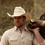 Bearded man wearing a 10X Cowboy Santos tan and ivory cowboy hat with plaid white shirt holding a brown horse saddle over his left shoulder