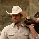 Bearded man wearing a 10X Cowboy Santos tan and ivory cowboy hat with plaid white shirt holding a brown horse saddle over his left shoulder