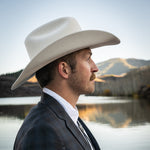 Man wearing a cowboy hat and suit standing by a lake with mountains in the background