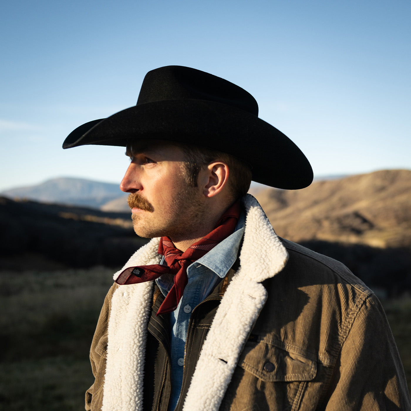 Man wearing a cowboy hat and brown jacket with a red bandana, standing in a desert mountain landscape.
