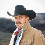 Man wearing a black cowboy hat and beige jacket with a red bandana, standing outdoors.